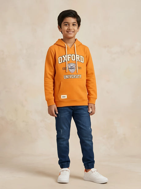 Young boy wearing an orange long-sleeve hooded t-shirt with Oxford University graphic print and kangaroo pocket paired with blue jeans.