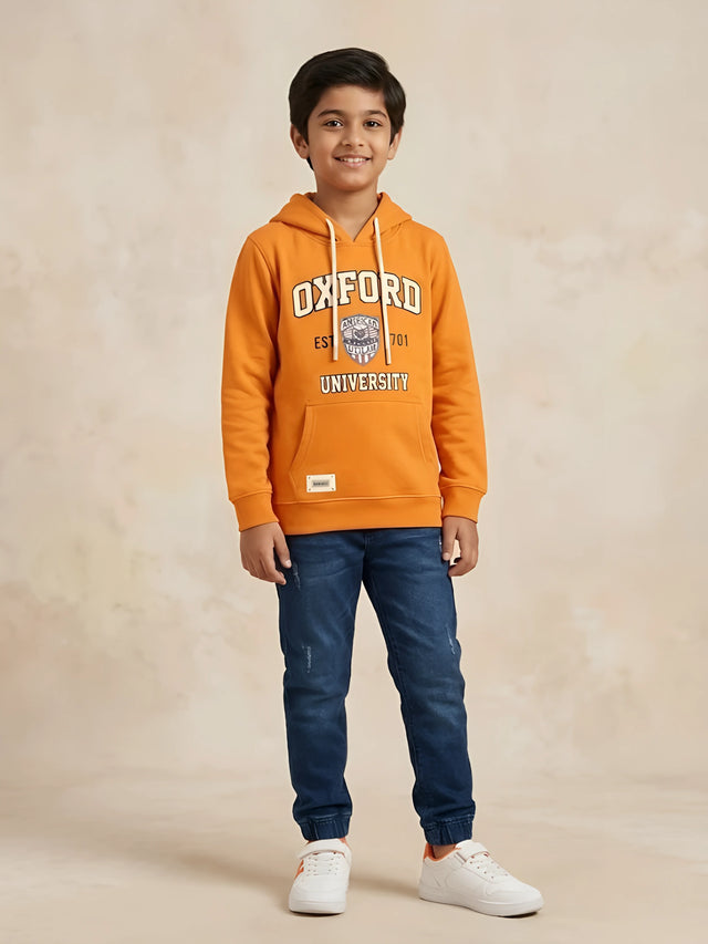 Young boy wearing an orange long-sleeve hooded t-shirt with Oxford University graphic print and kangaroo pocket paired with blue jeans.