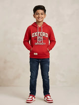 Young boy wearing a red hooded casual t-shirt with Oxford graphic print, white drawstrings, and a front kangaroo pocket paired with dark blue jeans