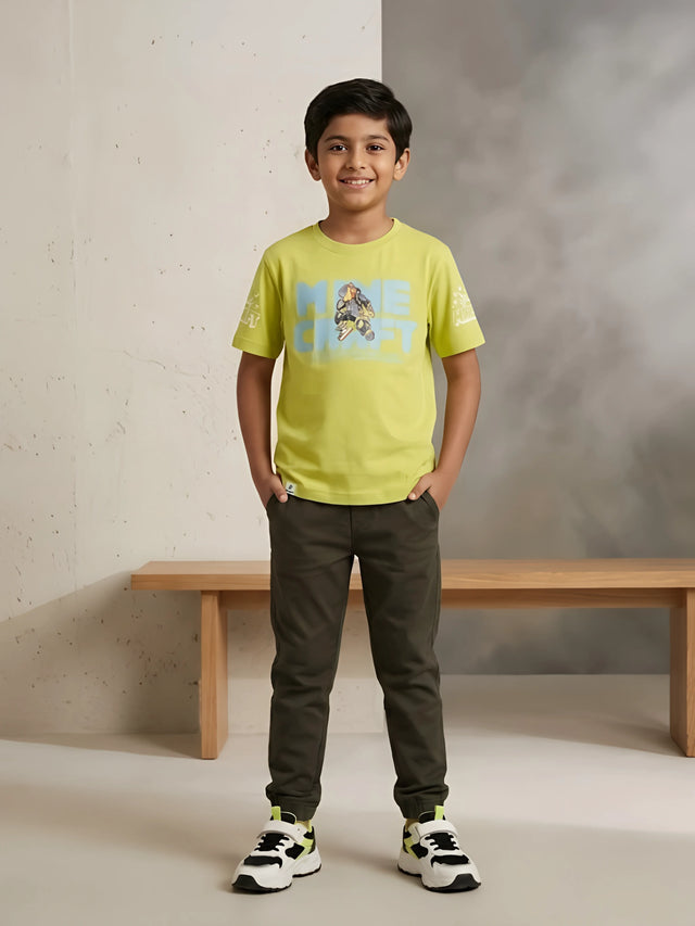 Young boy wearing a vibrant lime green short-sleeve casual graphic t-shirt paired with olive green trousers and sneakers.