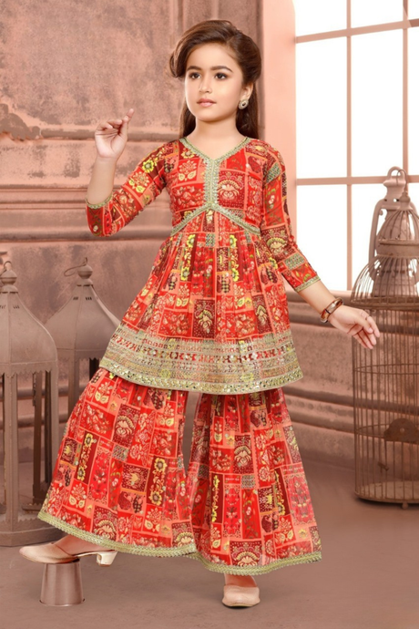 Young girl wearing a three-quarter sleeve red and orange printed sharara suit, featuring a flared kurti with decorative trim and voluminous sharara pants.