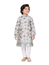 Full-length image of a young boy wearing a cream/off-white traditional kurta with all-over blue floral embroidery and a matching border, paired with white churidar pajamas and gold ethnic shoes.