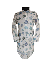 Full-length image of a young boy wearing a cream/off-white traditional kurta with all-over blue floral embroidery and a matching border, paired with white churidar pajamas and gold ethnic shoes.