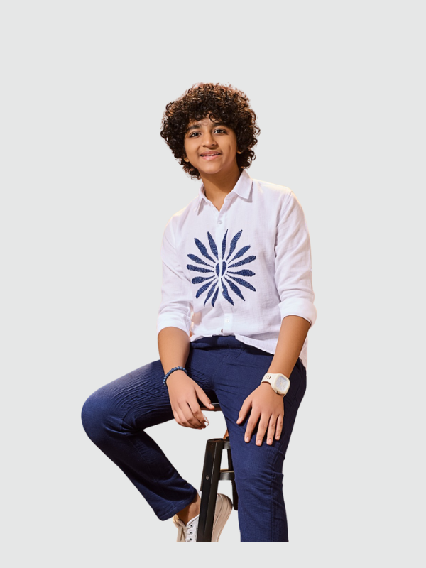Smiling older boy wearing a long-sleeve white shirt with a large navy blue sunburst print on the chest, paired with navy blue textured trousers and white sneakers. The outfit is a smart-casual coordinate set