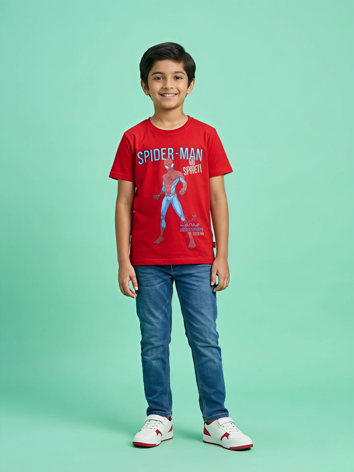 A smiling young boy wearing a bright red short-sleeve T-shirt with a Spider-Man graphic print on the front, paired with blue jeans and white sneakers, standing against a light green background.