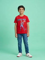 A smiling young boy wearing a bright red short-sleeve T-shirt with a Spider-Man graphic print on the front, paired with blue jeans and white sneakers, standing against a light green background.