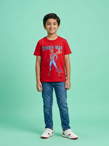 A smiling young boy wearing a bright red short-sleeve T-shirt with a Spider-Man graphic print on the front, paired with blue jeans and white sneakers, standing against a light green background.