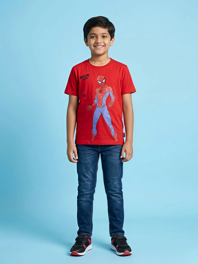 A young boy wearing a bright red short-sleeve crew neck t-shirt with a Spiderman graphic print on the front, paired with blue jeans and sneakers.