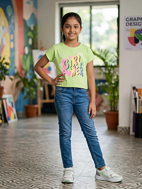 A young girl smiling and posing in a light green graphic t-shirt with 'Girl Power' printed on the front, paired with blue jeans and white sneakers.