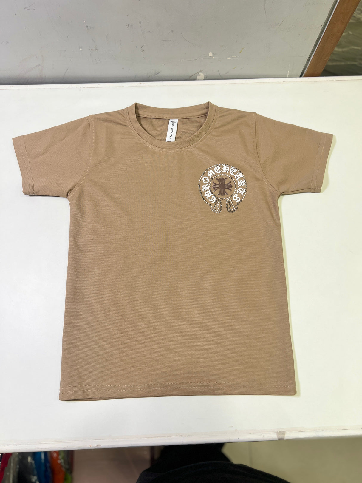 Tan/Brown boys' short-sleeve T-shirt with a small circular distressed cross and text logo graphic on the left chest, hanging on a white background.