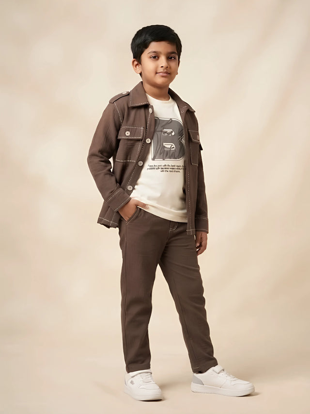 Young boy wearing a brown full sleeve casual shirt with contrast stitching over an off-white graphic t-shirt and matching brown trousers
