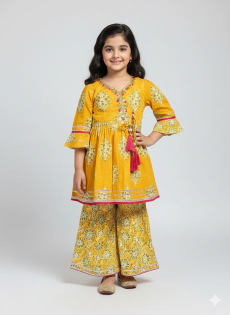 Young girl wearing a yellow peplum kurta with three-quarter bell sleeves and a pink-tasseled tie, matched with wide-leg printed sharara pants, posing against a grey background.
