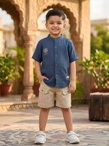 Young boy wearing a blue mandarin collar shirt with sun embroidery and khaki cargo shorts coordinate set standing in a courtyard.