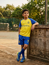 A young boy standing on an outdoor sports field wearing a yellow sports jersey with blue sleeves and matching blue shorts with the number 7.