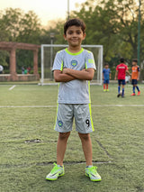 A young boy standing on a football field wearing a grey sports coordinate set with neon-green accents.