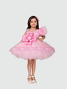 Young girl posing in a full, voluminous pink tulle party dress featuring a large, layered fabric bow on the bodice and a ruffle sleeve detail. She is wearing light pink formal shoes