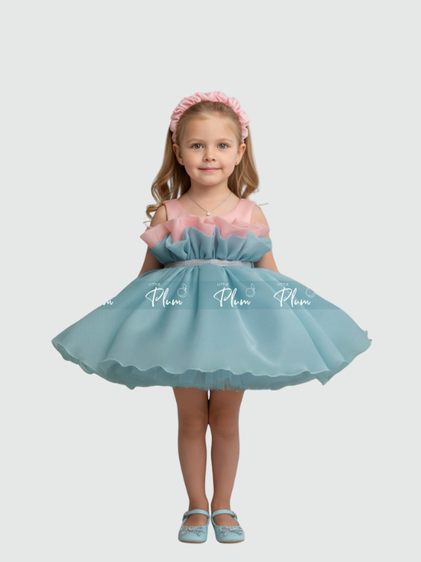 Young girl smiling in a two-tone color-block party dress with a blue skirt and pink ruffle neckline. The dress features a silver rhinestone belt and a voluminous tulle skirt, paired with a pink headband and blue shoes