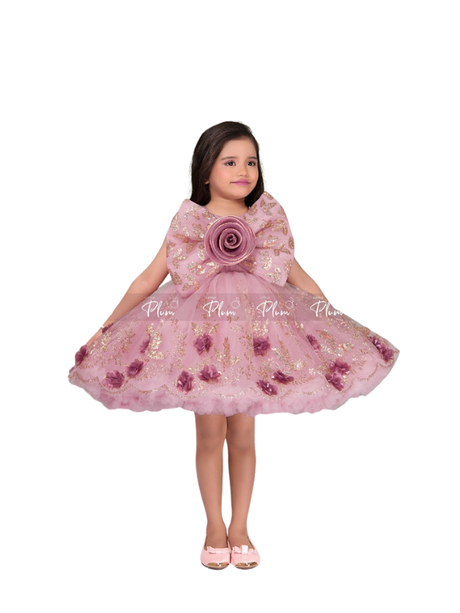 Full-length image of a young girl wearing a dusty rose pink party frock with a voluminous tulle skirt, all-over gold glitter, 3D mauve flowers, and a large matching bow on the chest.