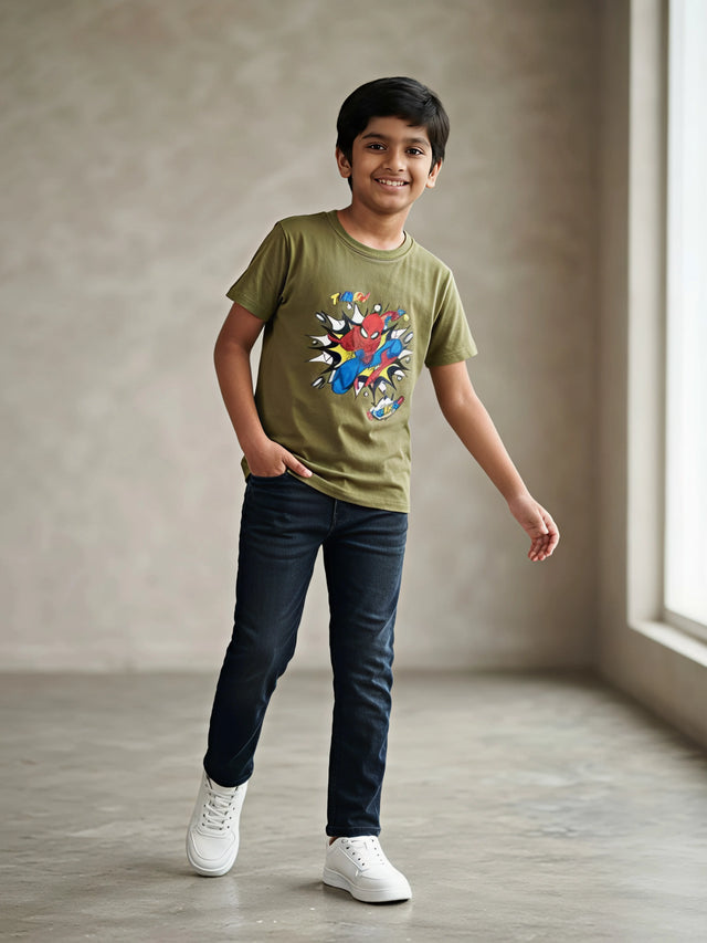 Young boy wearing an olive green round neck t-shirt with a Spider-Man superhero graphic print, paired with dark jeans and white sneakers.