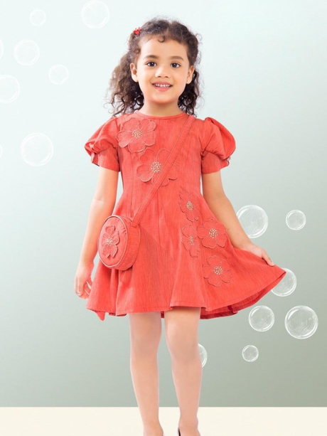 Young girl with curly hair wearing a rust orange colored frock with puff sleeves and 3D floral appliqués, carrying a matching round sling bag over her shoulder, standing amidst bubbles.