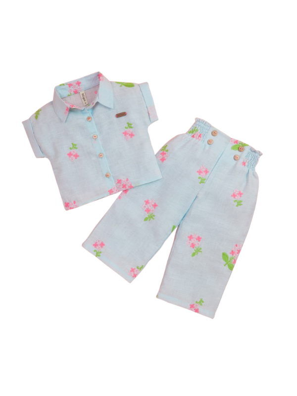 Girls' light blue short-sleeve collared shirt and matching wide-leg trousers set, both featuring a pink and green floral print. The trousers have a smocked waistband with wooden buttons.