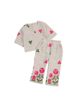 Girls' cream-colored two-piece set featuring a short-sleeve crop top and trousers, both decorated with pink and green floral appliqués. The trousers have large pink flower appliqués on the hem.