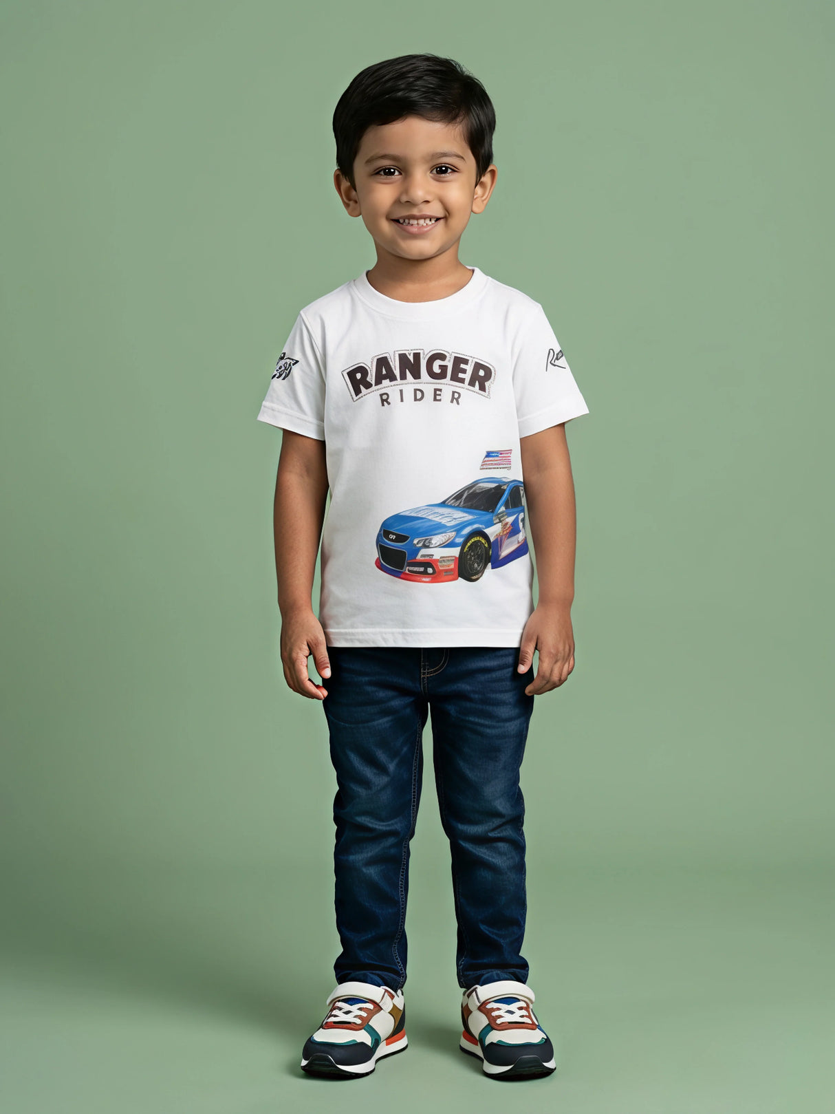 A young Indian boy smiling while wearing a white round-neck short-sleeve t-shirt featuring a blue racing car graphic and "RANGER RIDER" text printed on the front, styled with dark blue jeans and colorful sneakers.