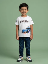 A young Indian boy smiling while wearing a white round-neck short-sleeve t-shirt featuring a blue racing car graphic and "RANGER RIDER" text printed on the front, styled with dark blue jeans and colorful sneakers.