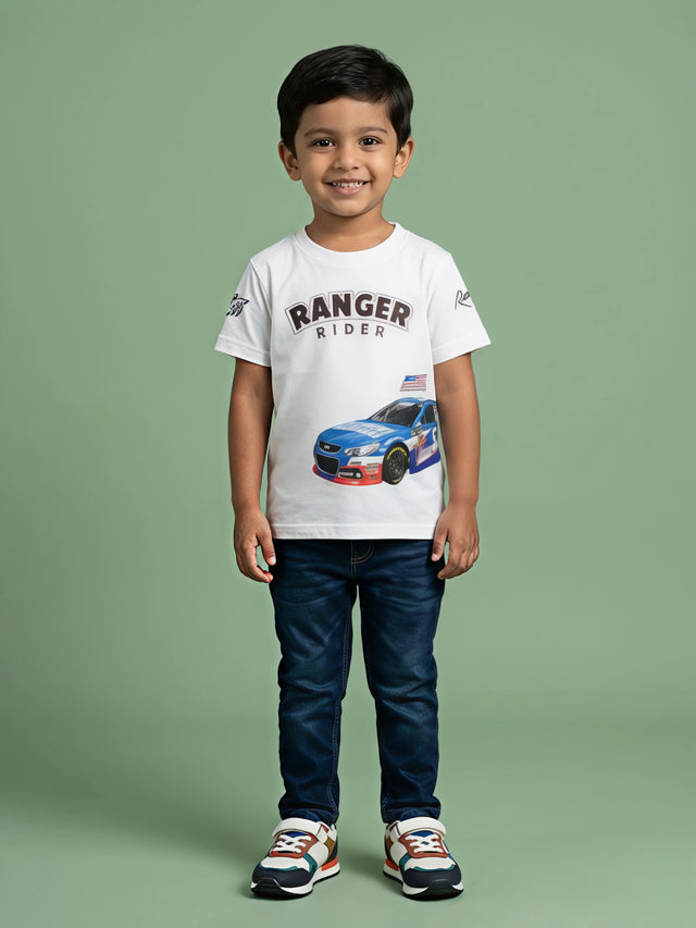 A young Indian boy smiling while wearing a white round-neck short-sleeve t-shirt featuring a blue racing car graphic and "RANGER RIDER" text printed on the front, styled with dark blue jeans and colorful sneakers.