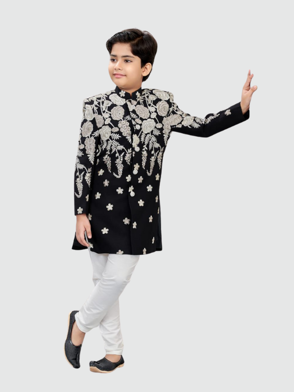 Young boy posing in a long, black Nehru-collar jacket with heavy silver/white floral embroidery, paired with crisp white trousers and black traditional footwear.