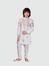 Young boy posing in a full-sleeve cream/off-white sherwani with all-over sequins and pastel floral embroidery, paired with light cream trousers and a long, multi-strand pearl necklace