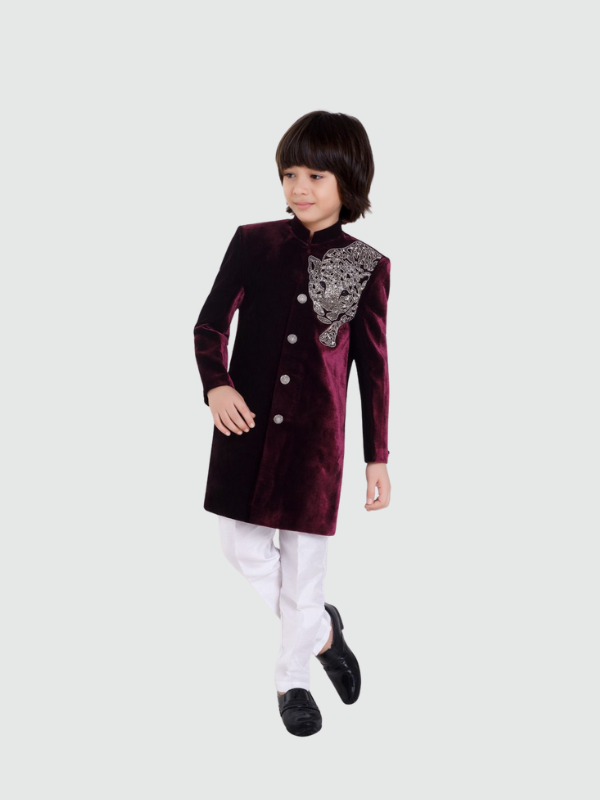Young boy posing in a full-sleeve burgundy/deep purple velvet sherwani with an elaborate silver metallic design on the shoulder, paired with white trousers and black formal shoes