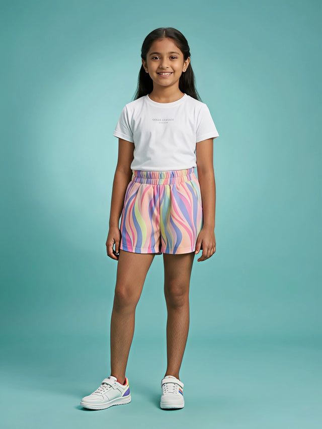 A young girl standing against a blue background wearing white sneakers, a white t-shirt, and pink casual shorts featuring a colorful wavy swirl pattern with a gathered elastic waistband, from the file TS15 - PINK - DAILY WEAR-artguru.jpg.