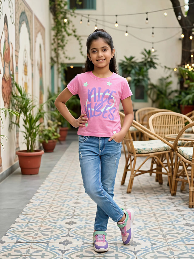 A happy young girl wearing a pink "Nice Vibes" graphic casual top paired with blue jeans.