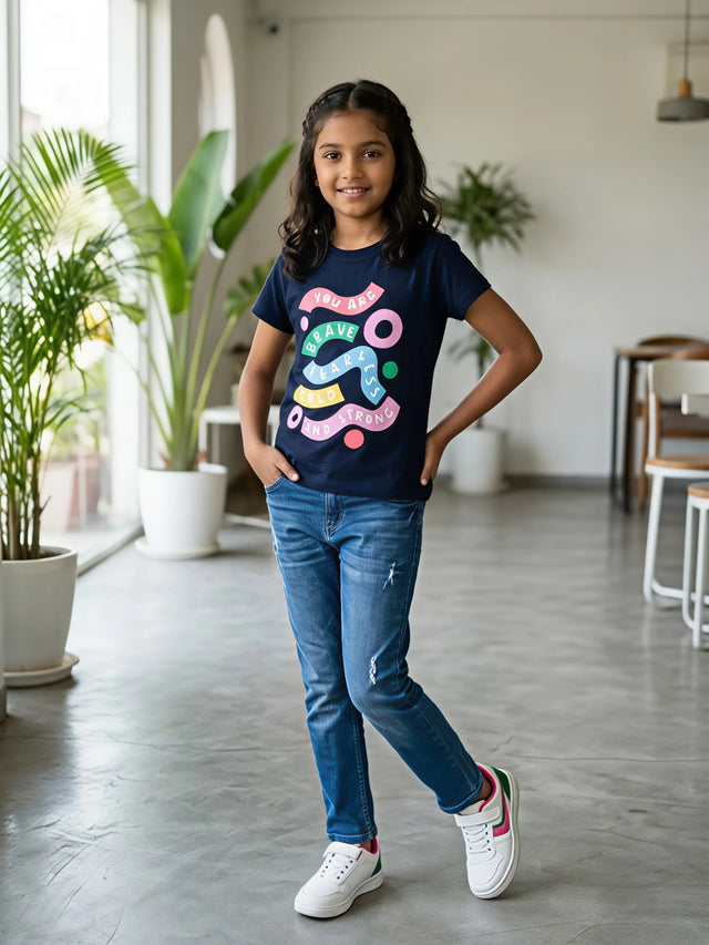 A young girl wearing a navy blue crew neck top with a colorful 'You Are Brave Fearless Bold and Strong' graphic print, paired with blue jeans.