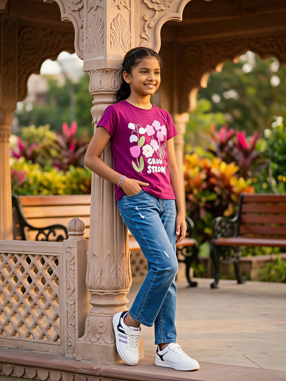 A young girl wearing a purple floral graphic top with "Grow Strong" text, paired with blue jeans and white sneakers in a park setting.