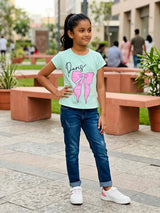 A young girl wearing a mint green short-sleeve top with a Paris and pink bow graphic print, styled with blue jeans.