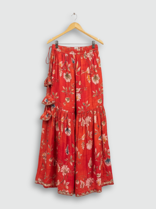 Full set laid flat: an orange/red floral-printed kurta with gold embroidered neckline, a matching tiered maxi skirt with tassels, and a printed dupatta with tassels.