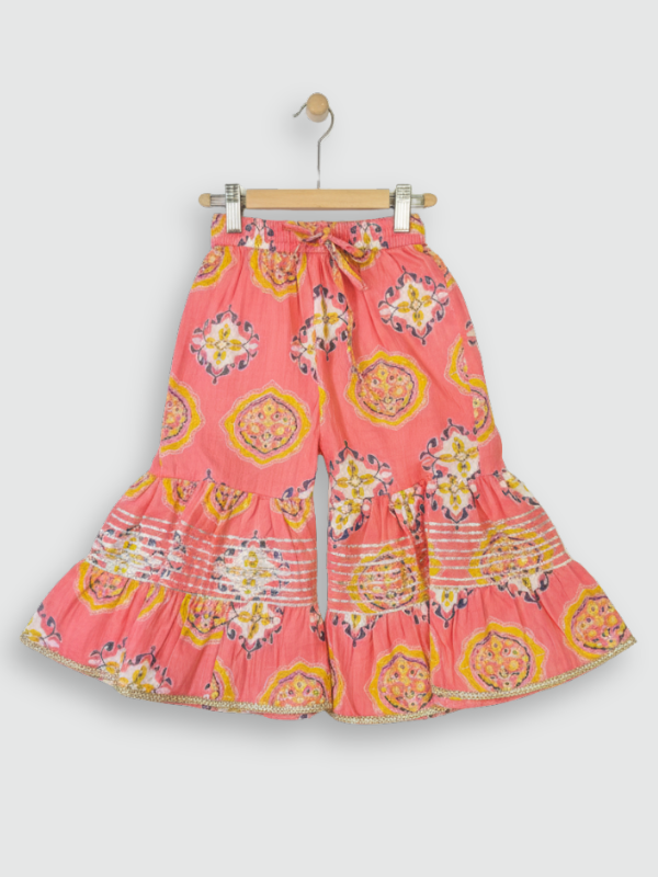 Full set laid flat: a coral pink Angrakha-style kurta with yellow/gold print and multi-colored pompoms, and matching tiered sharara pants with the same geometric print.