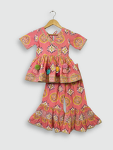 Full set laid flat: a coral pink Angrakha-style kurta with yellow/gold print and multi-colored pompoms, and matching tiered sharara pants with the same geometric print.