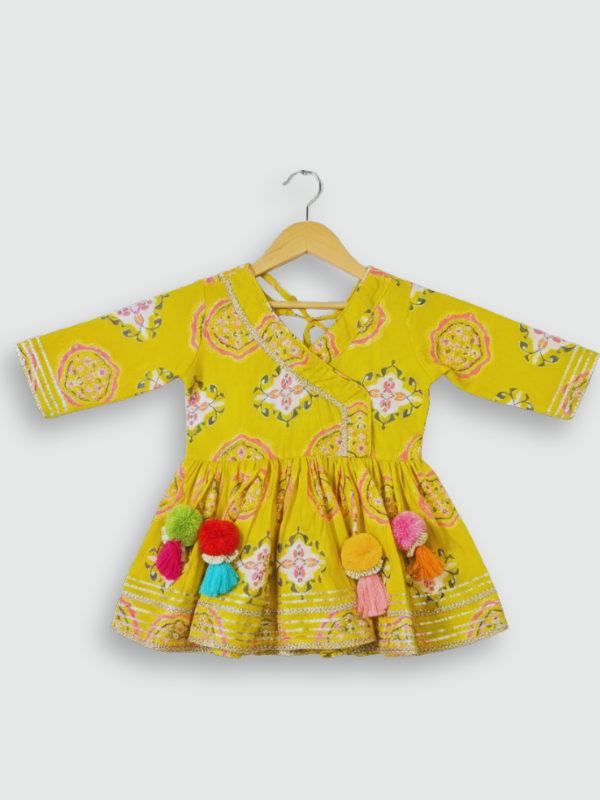 Full set laid flat: a mustard yellow Angrakha-style kurta with pink/white geometric print and multi-colored pompoms, and matching tiered sharara pants.