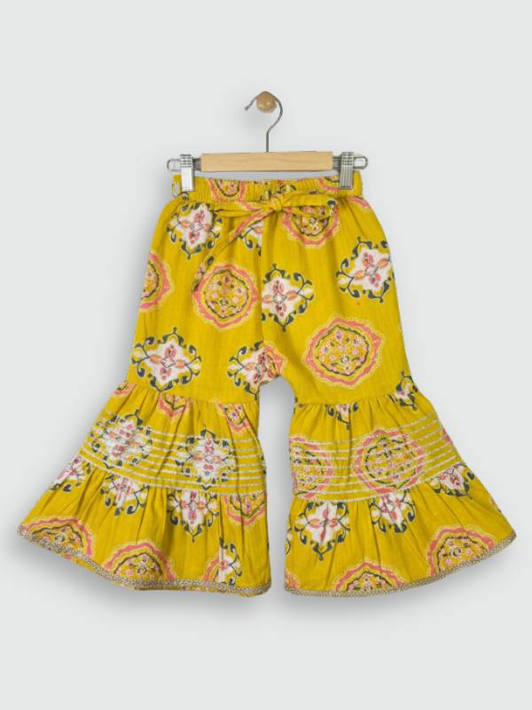 Full set laid flat: a mustard yellow Angrakha-style kurta with pink/white geometric print and multi-colored pompoms, and matching tiered sharara pants.