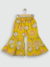 Full set laid flat: a mustard yellow Angrakha-style kurta with pink/white geometric print and multi-colored pompoms, and matching tiered sharara pants.