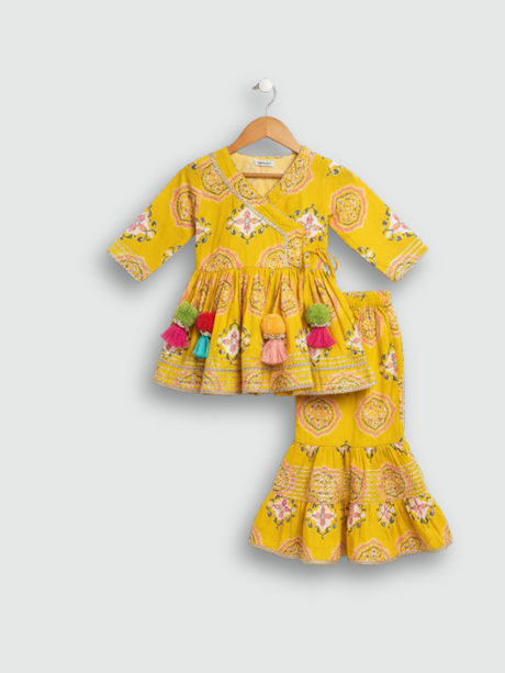 Full set laid flat: a mustard yellow Angrakha-style kurta with pink/white geometric print and multi-colored pompoms, and matching tiered sharara pants.