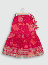 Full-length image of a young girl wearing a vibrant rani pink floral-printed ethnic set with an embroidered sleeveless peplum top, tiered sharara pants, and a pink-to-green ombre dupatta.