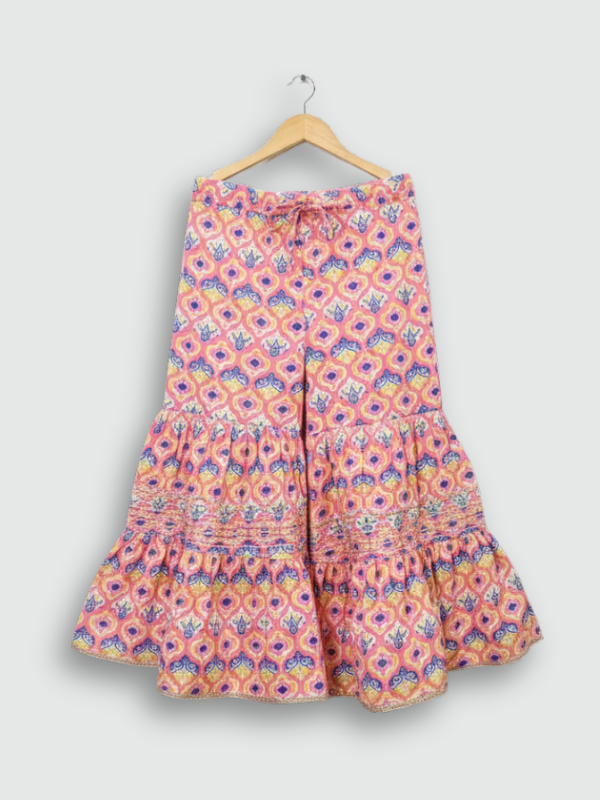 Full set laid flat: a coral pink Angrakha-style kurta with yellow/gold print and multi-colored pompoms, and matching tiered sharara pants with the same geometric print.