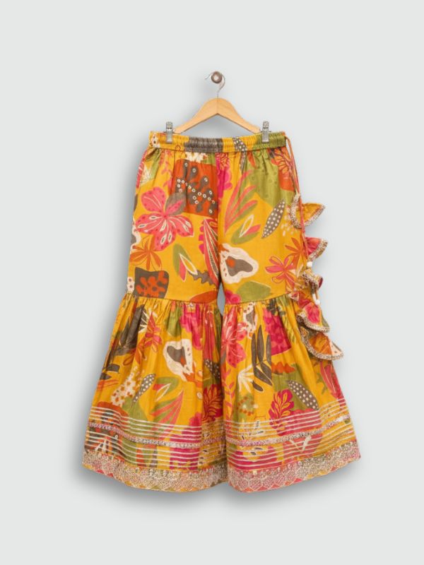 Full set laid flat: a mustard yellow Angrakha-style kurta with multicolor floral print and gold embroidery, tiered ruffle sharara pants, and a coordinating printed dupatta with tassels.