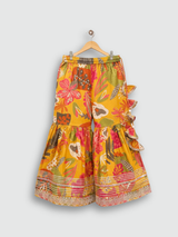 Full set laid flat: a mustard yellow Angrakha-style kurta with multicolor floral print and gold embroidery, tiered ruffle sharara pants, and a coordinating printed dupatta with tassels.