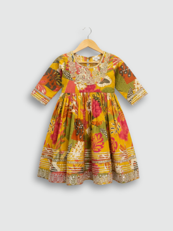 Full set laid flat: a mustard yellow Angrakha-style kurta with multicolor floral print and gold embroidery, tiered ruffle sharara pants, and a coordinating printed dupatta with tassels.