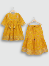 Full set laid flat: a mustard yellow Angrakha-style kurta with pink/white geometric print and multi-colored pompoms, and matching tiered sharara pants.